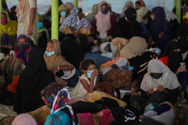 A Rohingya refugee child sits on the floor wearing a surgical face mask surroundedtightly  by other people, mostly women wearing head scarves in a large room.