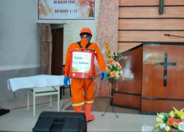A worker dressed in orange sprays disinfectant from a container which is labeled 'lazismu' inside a church