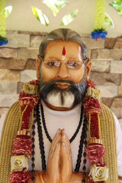 Photo of a realistic statue portraying an elderly man wearing rosaries and a flower garland. This was a late head pujari (priest) in a Guyanese Hindu temple, his hands folded to the Namaste greeting. Guyana, September 2017.