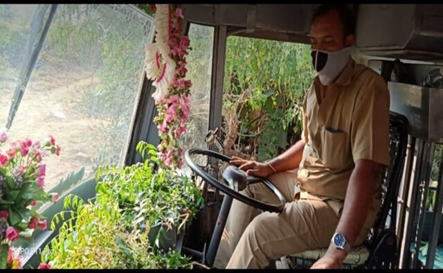 Photo of a man  wearing a mask and driving a bus decked with neem leaves.
