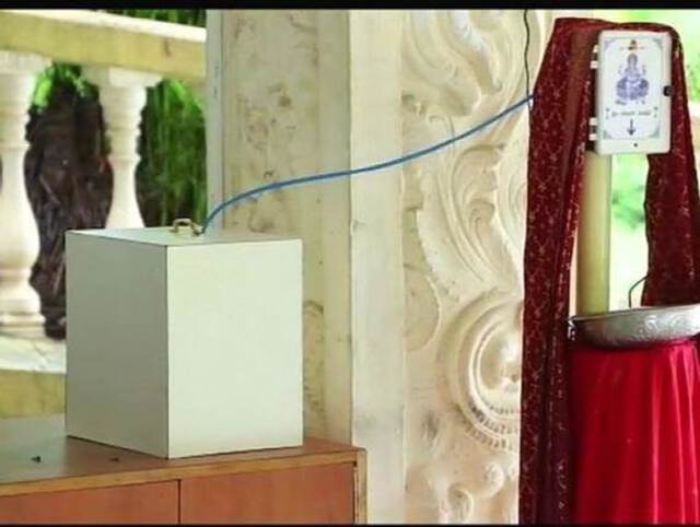 Photo of a box connected to a cable are seen in front of a white temple column. This is a mechanical holy water (water from river Ganga) dispenser installed in a Hindu Temple.