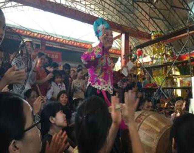 A photos of a male spirt-medium dressed in women's clothing dancing and smiling in the center of a a crowd of worshippers.