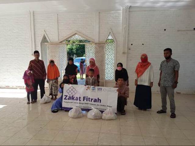 Men, women, and children stand behind a banner which reads "Zakat Fitrah" with plastic bags set in front of the banner