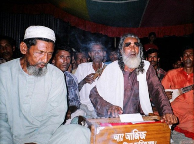 Photo of a man singing and playing a keyboard instrument, surrounded by others accompanying him.