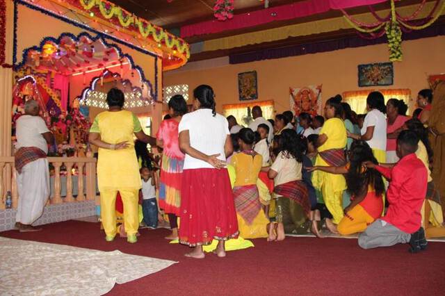 Photo of Hindu devotees in a temple, touching each other's shoulders while offerings are made to a deity. Guyana, September 2017.