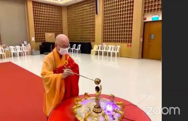 Photo of a male monk, wearing a face mask, in an empty hall. He is  bathing a statue of the Buddha. This is SBF’s President Venerable Seck Kwang Phing conducting the Bathing of the Buddha ritual via Zoom.